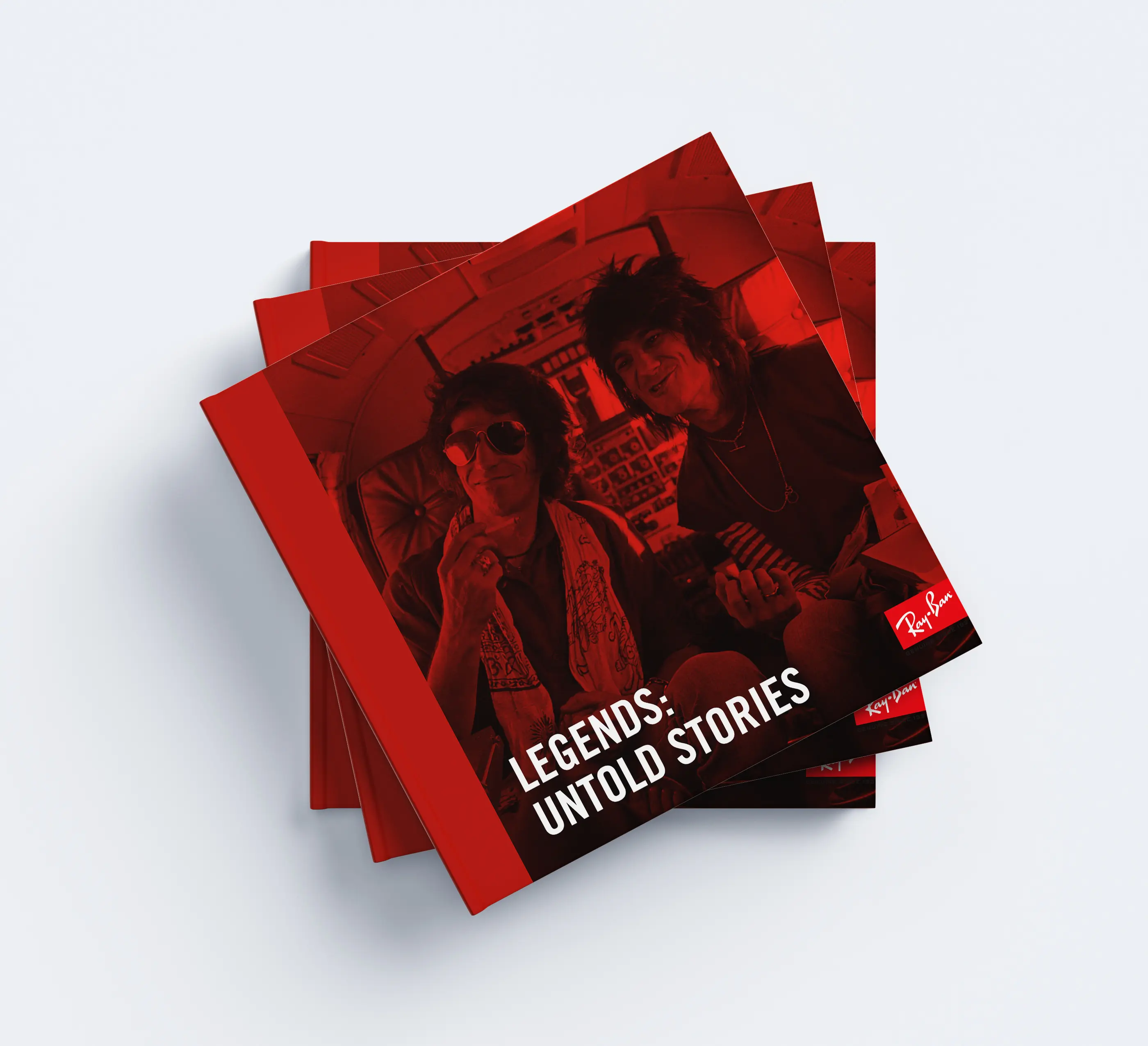 A stack of square red books titled Legends: Untold Stories featuring two people with long hair and sunglasses smiling on the cover. The Ray-Ban logo appears in the bottom right corner.